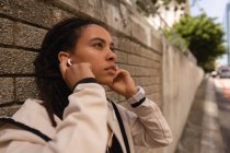 Seitenansicht einer jungen Frau mit gemischter Rasse, die Musik über Kopfhörer hört, während sie sich auf der Straße an eine Wand lehnt — Stockfoto
