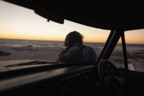 Вид сбоку романтической пары, стоящей у машины на пляже на закате — стоковое фото