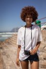Vista frontale di felice bella donna di razza mista in piedi con fotocamera in spiaggia in una giornata di sole — Foto stock