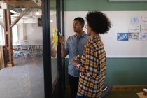 Вид збоку на молодих бізнесменів змішаної раси, які пишуть на липких нотах, що стоять в сучасному офісі — стокове фото