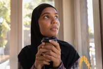 Vista frontal da jovem pensativa no hijab tomando café em um café — Fotografia de Stock