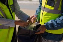 Parte média de voluntários limpando praia em um dia ensolarado. macho é segurando saco de plástico enquanto fêmea coloca o lixo no saco . — Fotografia de Stock