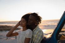 Вид сбоку романтической афроамериканской пары, склоняющейся к машине на пляже на закате — стоковое фото