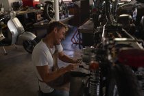 Seitenansicht eines kaukasischen Fahrradmechanikers, der beim Reparieren eines Fahrrads in der Garage zum Handy greift — Stockfoto