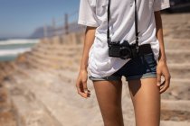 Средняя часть женщины, стоящей с камерой на пляже в солнечный день — стоковое фото