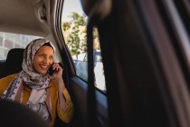 Vista frontal de feliz hermosa mujer de raza mixta sonriendo y hablando por teléfono móvil mientras viaja en coche - foto de stock