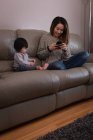 Front view of a happy Asian mother and daughter using mobile phone while sitting on sofa at home — стокове фото