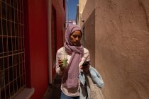 Vista frontal de la hermosa mujer de raza mixta utilizando el teléfono móvil mientras camina y sostiene batido en el callejón - foto de stock