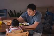 Вид спереди на отца-азиата, кормящего свою дочь за обеденным столом на кухне дома — стоковое фото