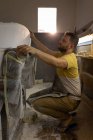 Vue latérale de l'homme caucasien concentré vérifiant la forme des planches de surf dans un atelier. Il est en position accroupie. . — Photo de stock