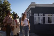 Вид спереду змішаної раси друзі-жінки тримають каву в очікуванні перетину дороги на вулиці в сонячний день — стокове фото