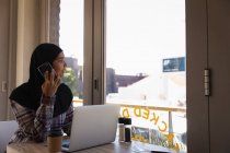 Vista lateral da jovem mulher no hijab falando no telefone celular enquanto usa laptop em um café, enquanto olha para fora — Fotografia de Stock