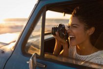 Вид сбоку счастливой красивой афроамериканки, делающей фотографии цифровой камерой, сидя в машине на пляже на закате — стоковое фото