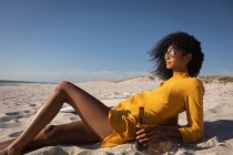 Вид сбоку красивой молодой афроамериканки, склоняющейся на пляже во время питья в солнечный день — стоковое фото