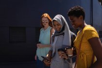 Vista lateral de jóvenes amigas de raza mixta que interactúan entre sí mientras caminan por la calle de la ciudad - foto de stock