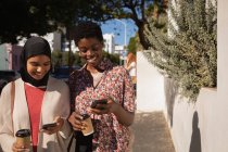 Vista frontal da raça mista feliz amigos do sexo feminino usando telefone celular enquanto toma café na rua no dia ensolarado — Fotografia de Stock