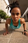 Retrato de una joven mujer de raza mixta escuchando música en los auriculares mientras hace ejercicio push-up en la ciudad - foto de stock