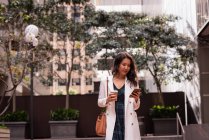 Vista frontal de la mujer asiática usando el teléfono móvil mientras está de pie en la calle - foto de stock