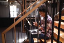 Вид збоку красива бізнес-леді змішаної раси, що використовує мобільний телефон, сидячи на сходах з ноутбуком — стокове фото