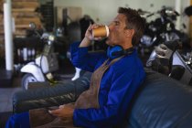 Seitenansicht eines kaukasischen Fahrradmechanikers, der Kaffee trinkt, während er es sich auf dem blauen Sofa in der Garage gemütlich macht — Stockfoto