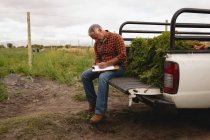 Side view of Caucasian male farmer maintaining the record of plants on clipboard while sitting on backside of car — Stock Photo