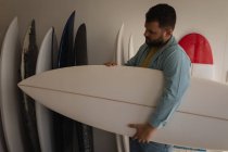 Vista lateral do homem caucasiano segurando nova prancha de surf enquanto olha para a sua criação em uma oficina — Fotografia de Stock