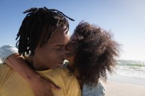 Vista frontal de la joven pareja afroamericana romántica de pie con el brazo alrededor en la playa en el día soleado - foto de stock
