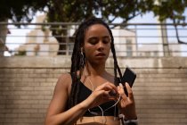 Вид спереду молодої змішаної раси жінка в навушниках при використанні мобільного телефону на вулиці міста — стокове фото