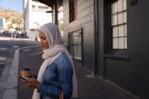 Vista laterale di una donna di razza mista che usa il telefono cellulare e tiene un caffè in piedi sul marciapiede — Foto stock