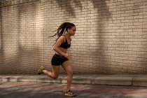 Vue latérale de la jeune femme de race mixte écoutant de la musique sur des écouteurs tout en faisant du jogging dans la rue — Photo de stock