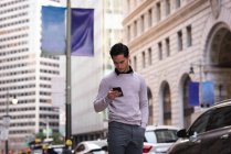 Vorderansicht eines hübschen jungen asiatischen Mannes mit Handy, während er auf der Straße steht — Stockfoto