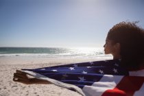 Vista posteriore della giovane donna afroamericana con bandiera americana in piedi sulla spiaggia in una giornata di sole — Foto stock