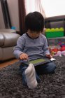 Vista frontal de un niño asiático pequeño usando una tableta digital mientras está sentado en la alfombra en casa - foto de stock