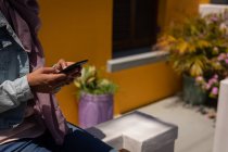 Partie médiane de la femme utilisant un téléphone portable tout en étant assis sur le mur en face de la maison — Photo de stock