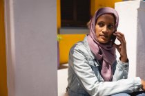Vista frontal de la hermosa mujer de raza mixta reflexiva con hijab sentado frente a su casa mientras habla por teléfono móvil - foto de stock