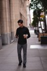 Vista frontale di bell'uomo asiatico che utilizza il telefono cellulare con caffè mentre si cammina dalla strada — Foto stock