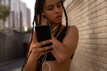 Vista frontal de la joven mujer de raza mixta que usa auriculares mientras usa el teléfono móvil en la calle de la ciudad - foto de stock