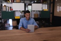 Вид спереду щасливого бізнесмена змішаної раси, використовуючи цифровий планшет за столом, сидячи в сучасному офісі — стокове фото