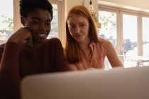 Вид спереду на молоду змішану расу друзі-жінки взаємодіють один з одним, використовуючи ноутбук в ресторані — стокове фото