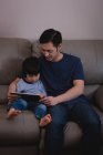Вид спереди отца-азиата и его сына, сидящих дома на диване с цифровым планшетом — стоковое фото
