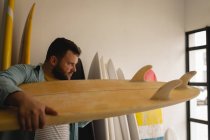Vista lateral del hombre caucásico revisando minuciosamente la tabla de surf en un taller - foto de stock