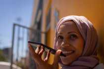 Крупным планом счастливой смешанной расы женщина улыбается и разговаривает по мобильному телефону, прислонившись к стене в солнечный день — стоковое фото