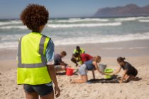 Vista posteriore della volontaria di razza mista in piedi sulla spiaggia mentre guarda gli altri volontari in una giornata di sole — Foto stock