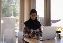 Vista frontal da jovem mulher no hijab usando o telefone celular enquanto trabalhava em um laptop em um café — Fotografia de Stock