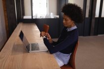 Vista lateral da jovem mulher de negócios mista verificando o tempo ao usar o laptop na mesa no escritório moderno — Fotografia de Stock
