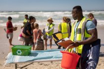 Vue de face du bénévole afro-américain debout avec seau et presse-papiers tandis que les autres bénévoles parlent derrière lui sur la plage — Photo de stock