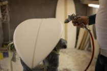 Partie médiane de l'homme peignant une planche de surf avec un pistolet à peinture en atelier — Photo de stock