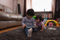Vue de face du frère et de la sœur asiatiques regardant ensemble la tablette numérique dans le salon à la maison — Photo de stock
