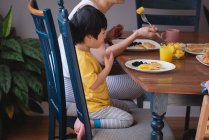 Vista lateral da mãe e do filho asiáticos desfrutando de café da manhã na mesa de jantar na cozinha em casa — Fotografia de Stock