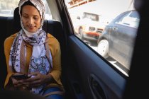 Вид женщины смешанной расы, улыбающейся и использующей мобильный телефон во время поездки в автомобиле в солнечный день — стоковое фото